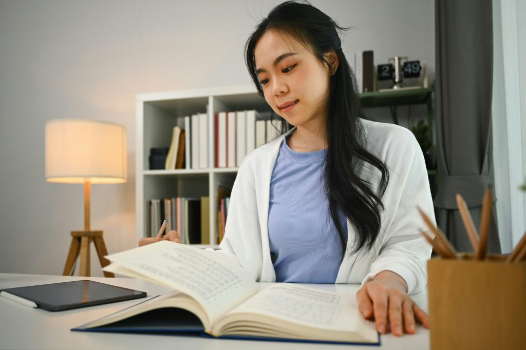 Charming asian fe male student reading book, preparing for exam or, doing homework in living room