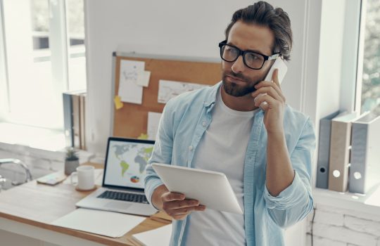 Confident man talking on phone and holding digital tablet while standing in the office