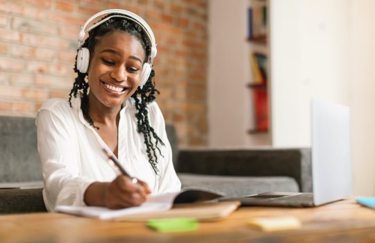 Happy black woman in headphones studing online with laptop from home, watching webinar and taking